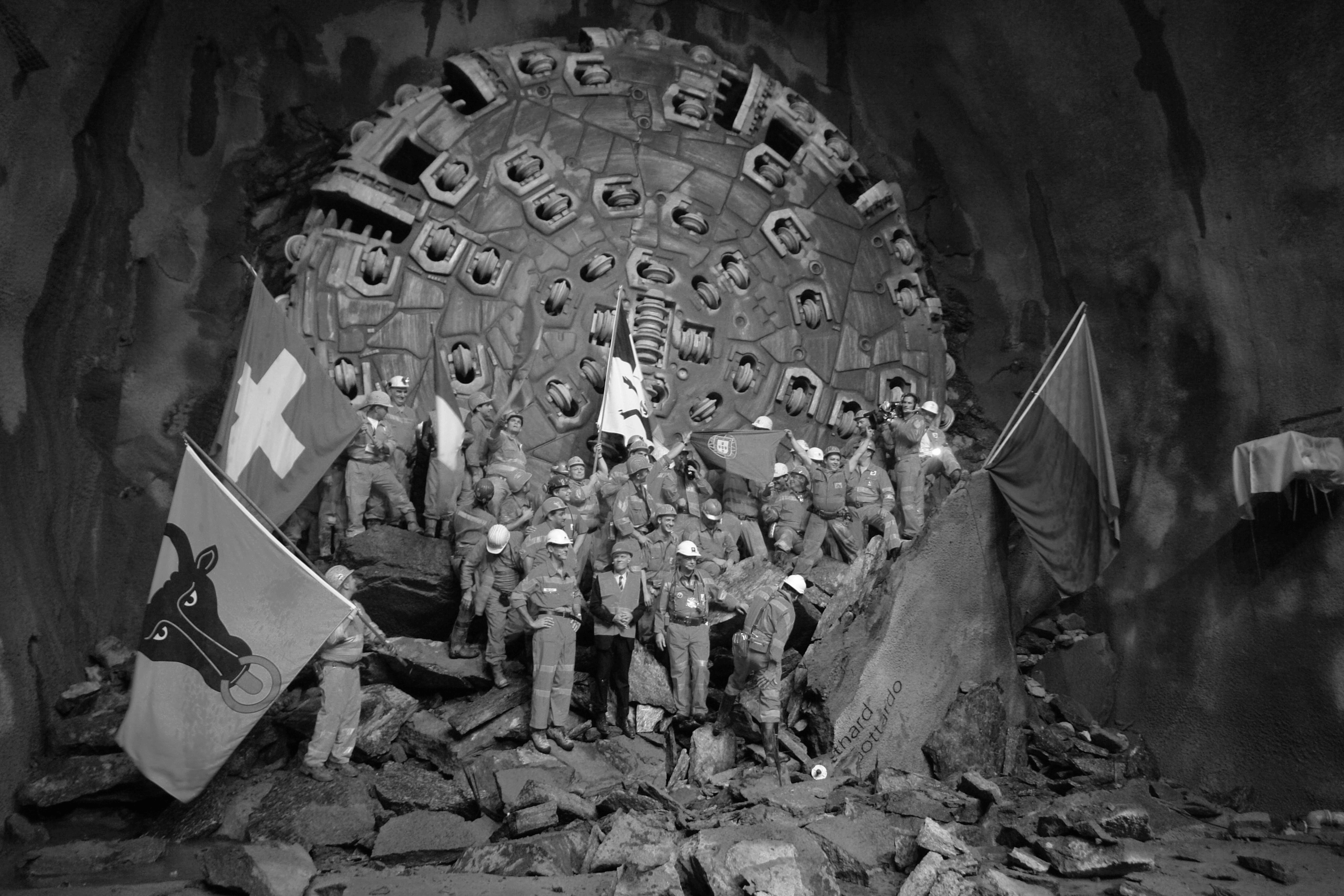 Schwarz-Weiß-Foto von Arbeitern, die vor einer großen Stahlbohrmaschine im Gotthardtunnel stehen und den Durchbruch feiern