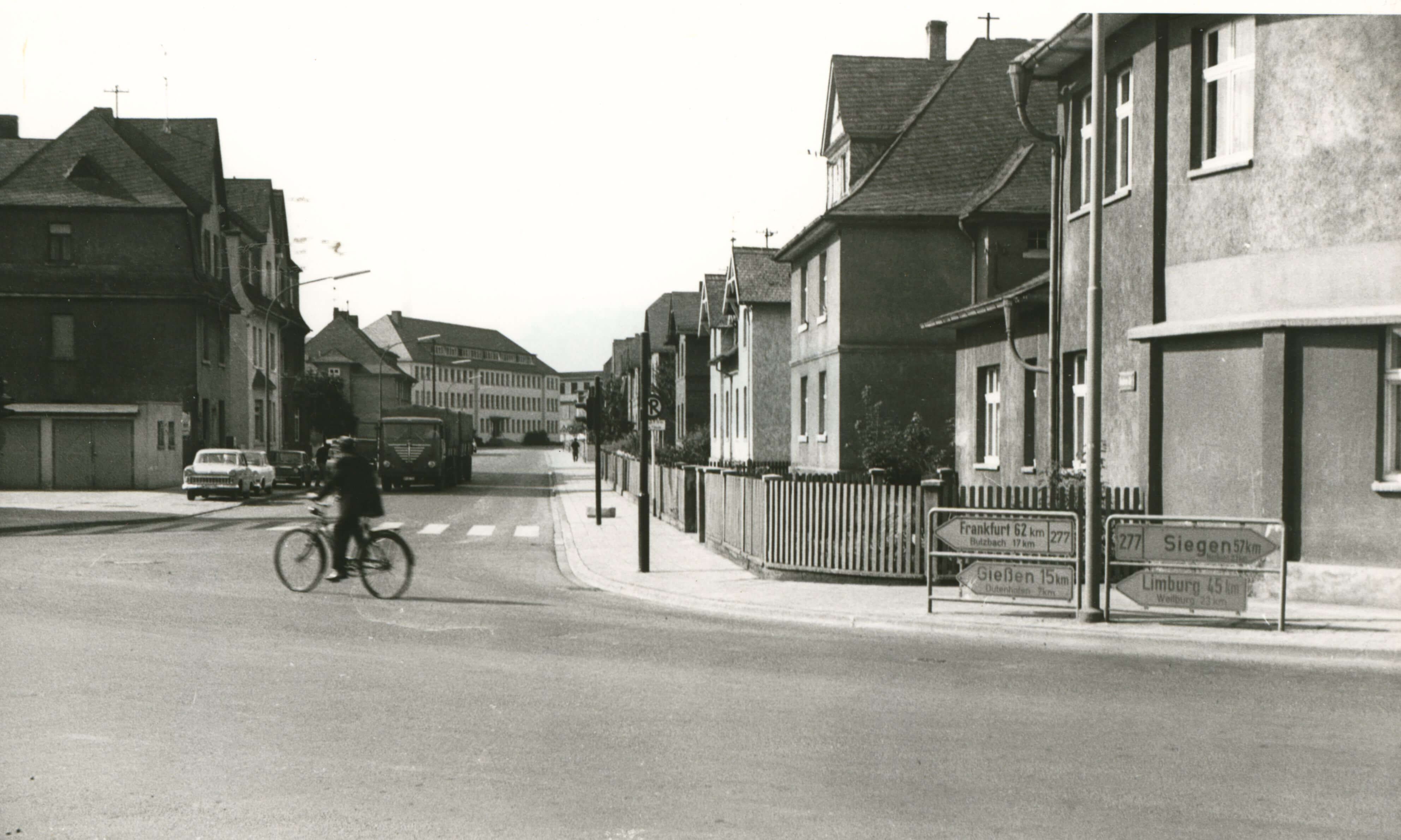 Ein historisches Schwarz-Weiß-Foto, das eine Straßenecke mit Häuserreihen und einem vorbeifahrenden Mann auf einem Fahrrad zeigt.