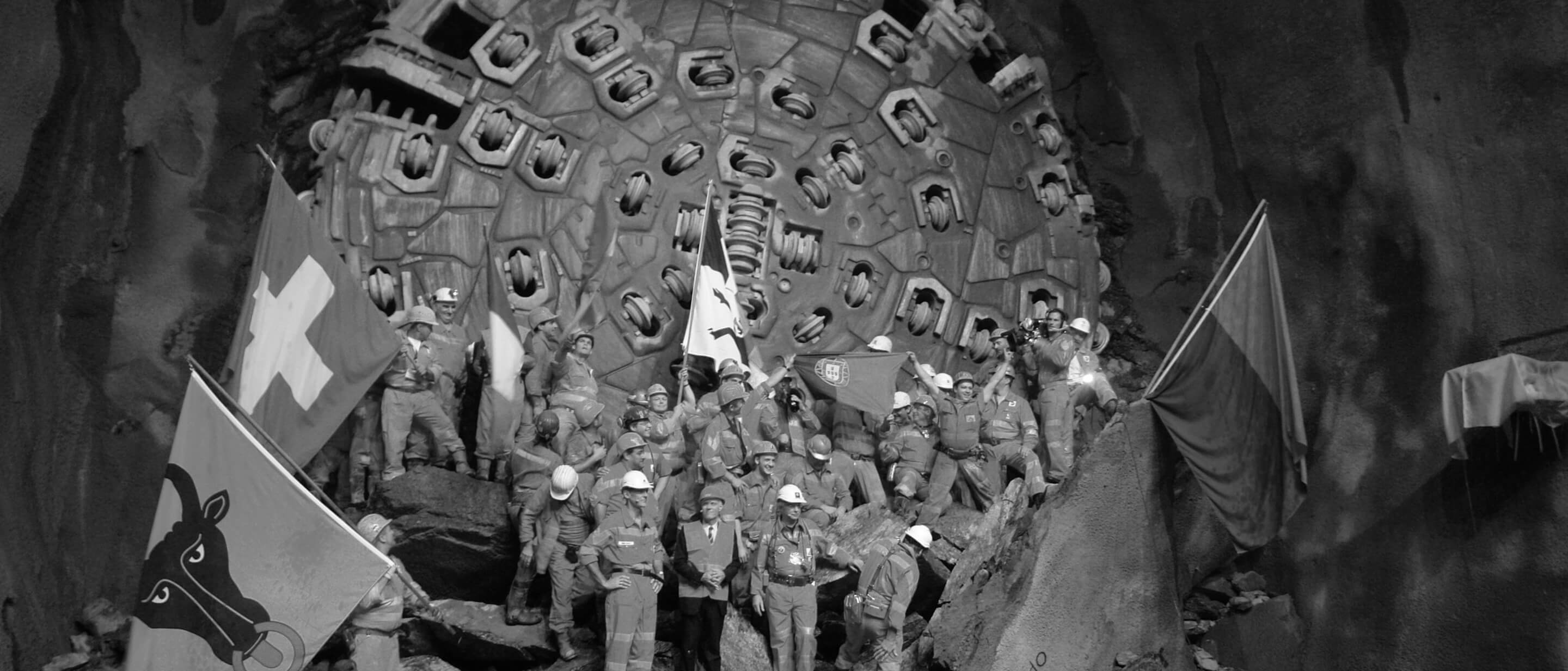 Schwarz-Weiß-Foto von Arbeitern, die vor einer großen Stahlbohrmaschine im Gotthardtunnel stehen und den Durchbruch feiern