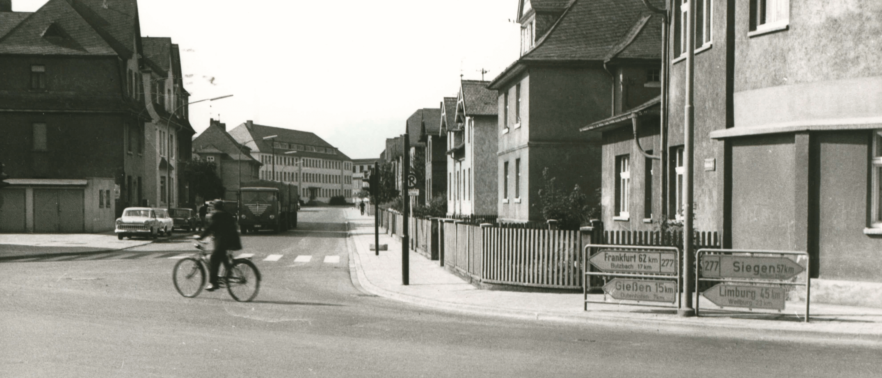 A historic photo in black and white depicting a street corner with rows of houses and a man driving past on a bike.